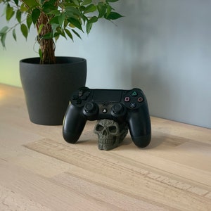 May include: A black PlayStation 4 controller resting on a green skull-shaped stand. The controller is on a light wood surface.