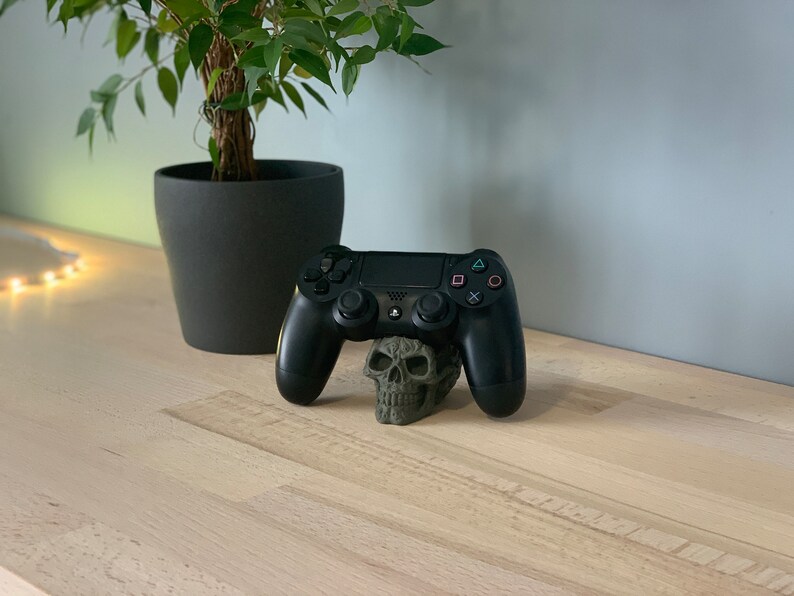 May include: A black PlayStation 4 controller resting on a green skull-shaped stand. The controller is on a light wood surface.