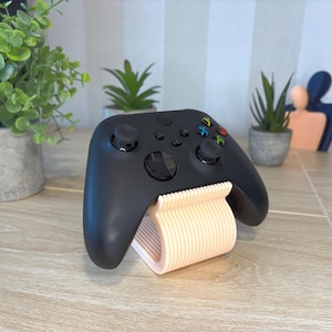 Op de afbeelding: Zwarte Xbox-controller op een lichtperzikstandaard. De controller heeft gekleurde knoppen en twee joysticks. De standaard heeft een gebogen ontwerp met verticale lijnen.