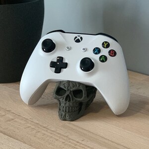 May include: A white Xbox controller is resting on a grey skull-shaped stand. The controller has black buttons and a black directional pad. The skull is detailed with intricate carvings.