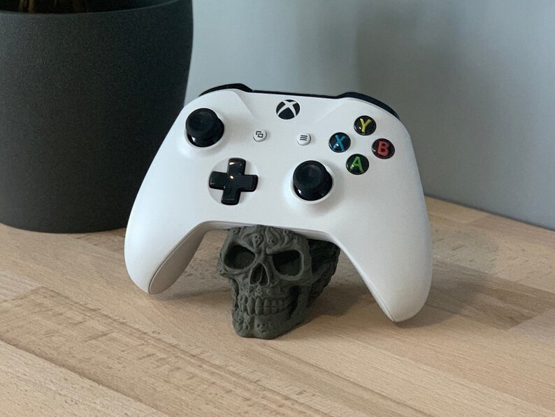 May include: A white Xbox controller is resting on a grey skull-shaped stand. The controller has black buttons and a black directional pad. The skull is detailed with intricate carvings.