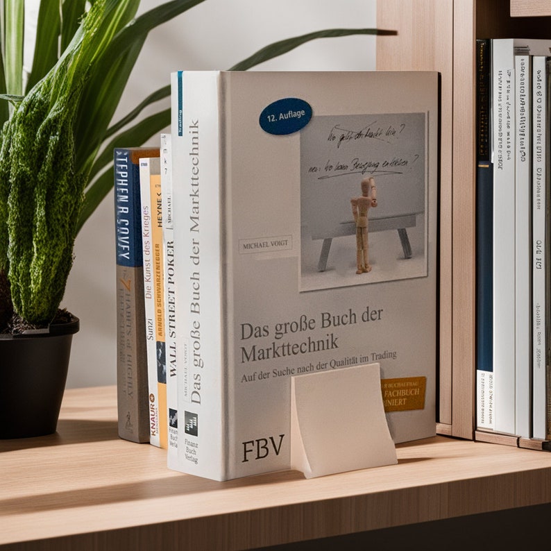 May include: A bookshelf with several books, including "The 7 Habits of Highly Effective People" by Stephen R. Covey, "The Art of War" by Sun Tzu, and "Wall Street Poker" by Michael Voigt. The book "Das gro&szlig;e Buch der Markttechnik" by Michael Voigt is prominently displayed on the shelf, with a white bookend in front of it.