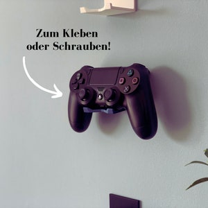 Op de afbeelding: Een zwarte PlayStation 4 controller gemonteerd aan een muur met de tekst "Zum Kleben oder Schrauben!" in wit erboven.