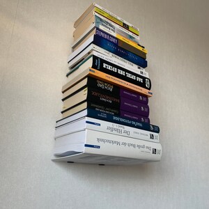 Peut inclure: Une pile de livres sur une &eacute;tag&egrave;re murale blanche. Les livres sont principalement en allemand et comprennent des titres tels que "Das Spiel der Spiele", "Rich Dad Poor Dad" et "Trading Psychology".