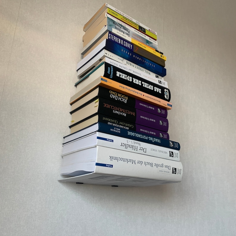 Peut inclure: Une pile de livres sur une &eacute;tag&egrave;re murale blanche. Les livres sont principalement en allemand et comprennent des titres tels que "Das Spiel der Spiele", "Rich Dad Poor Dad" et "Trading Psychology".