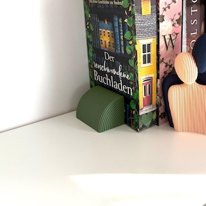 Peut inclure: Un serre-livres vert semi-circulaire avec un design strié soutient un livre intitulé "Der verschwundene Buchladen". Le livre présente une illustration d'une maison jaune. D'autres livres sont visibles en arrière-plan.