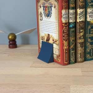 May include: A blue bookend with a curved design holds a row of seven hardcover books with gold lettering and illustrations. The books are titled "Harry Potter" in German and feature the titles "Und der Stein der Weisen", "Und die Kammer des Schreckens", "Gefangene von Askaban", "Der Feuerkelch", and "Der Halbblutprinz".