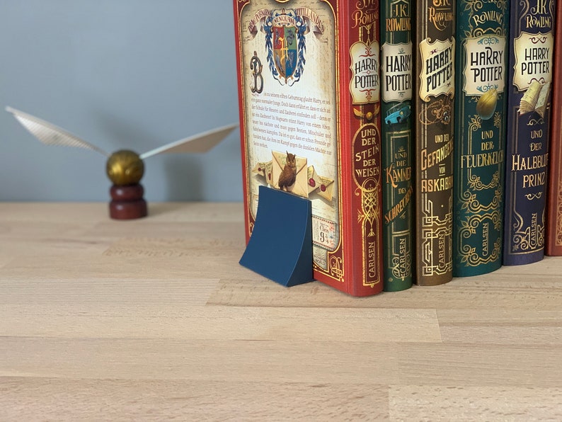 May include: A blue bookend with a curved design holds a row of seven hardcover books with gold lettering and illustrations. The books are titled "Harry Potter" in German and feature the titles "Und der Stein der Weisen", "Und die Kammer des Schreckens", "Gefangene von Askaban", "Der Feuerkelch", and "Der Halbblutprinz".