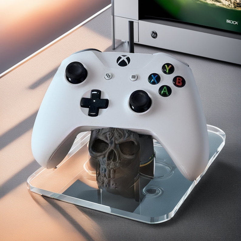 May include: A white Xbox controller with black buttons and a skull-shaped base. The controller is on a clear glass charging stand.