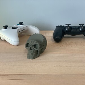 May include: A white and a black video game controller are placed on a wooden surface. A green skull-shaped object is in the centre of the controllers.