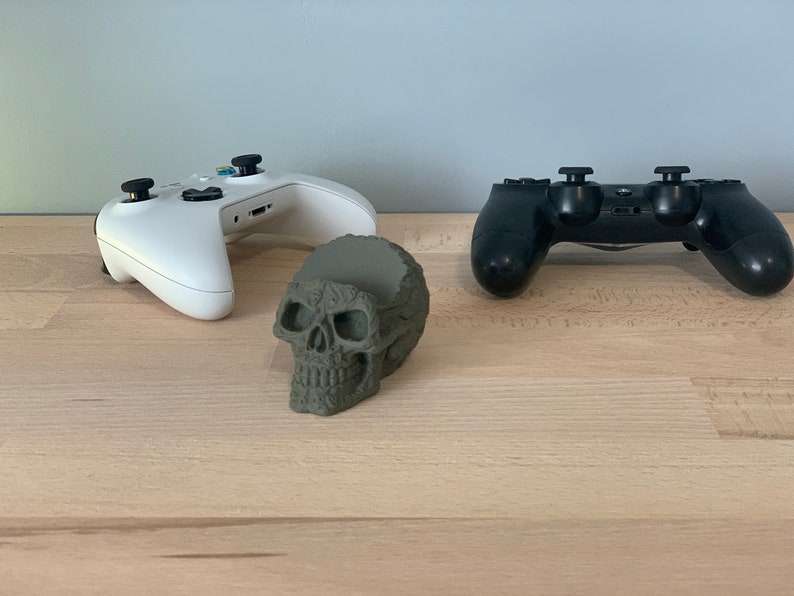 May include: A white and a black video game controller are placed on a wooden surface. A green skull-shaped object is in the centre of the controllers.