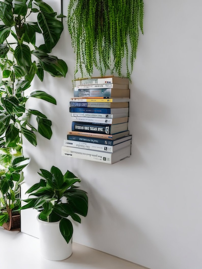 Op de afbeelding: Een stapel boeken op een witte wandplank met een groene plant die erboven hangt. De boeken zijn meestal blauw en hebben witte titels. Er staat een groene plant in een witte pot aan de linkerkant van de afbeelding.