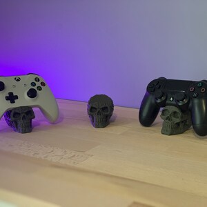 May include: Two video game controllers, one white and one black, are displayed on a wooden surface. Each controller is resting on a small, grey skull-shaped stand.