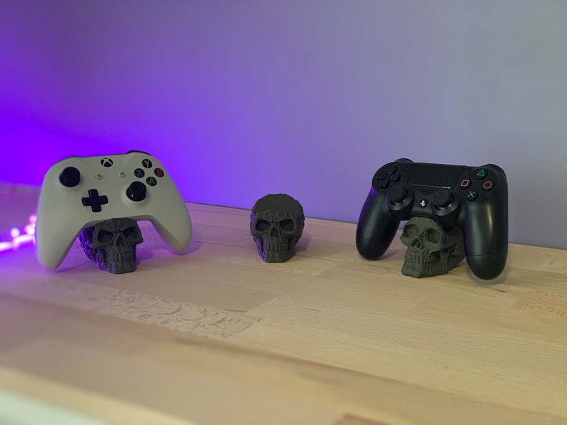 May include: Two video game controllers, one white and one black, are displayed on a wooden surface. Each controller is resting on a small, grey skull-shaped stand.