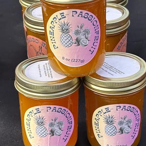 May include: Six glass jars filled with a bright orange liquid. The jars have gold lids and labels that say "Pineapple-Passionfruit" with a pineapple and passionfruit illustration. The labels also say "8 oz (227g)".