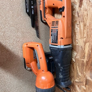 May include: Two orange and black power tools hanging on a wall. The larger tool is a reciprocating saw with a black blade. The smaller tool is a cordless flashlight. Both tools have the brand name "RIDGID" printed on them.