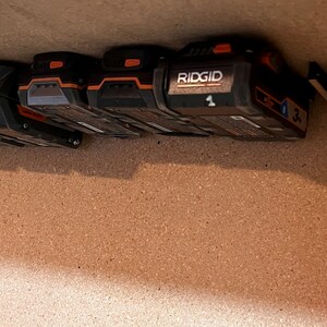 May include: A close-up of a wall-mounted battery storage system for power tools. The system features multiple black and orange batteries, including a RIDGID battery labeled with the number "1".