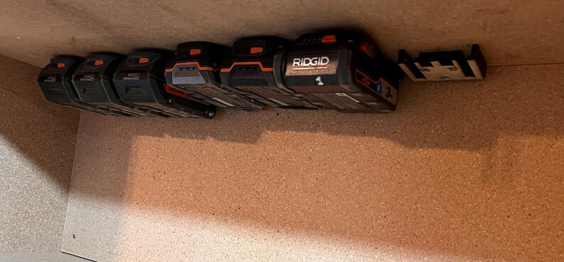 May include: A close-up of a wall-mounted battery storage system for power tools. The system features multiple black and orange batteries, including a RIDGID battery labeled with the number "1".