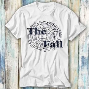 May include: White t-shirt featuring a circular black and white design with the words "The Fall" in dark blue. The design is on the front. The shirt has short sleeves and a crew neck.