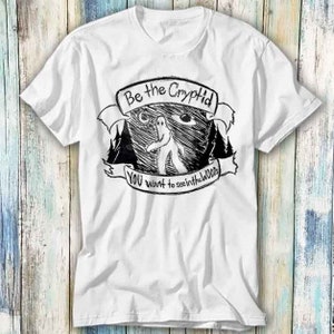 Puede incluir: Camiseta blanca con un estampado gráfico en blanco y negro de una criatura en el bosque. El texto de la camiseta dice "Be the Cryptid You want to see in the woods."