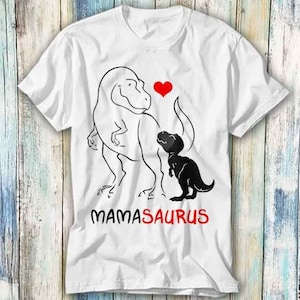 Puede incluir: Camiseta blanca con una ilustración en blanco y negro de dos dinosaurios. El dinosaurio más grande está delineado en negro y tiene un corazón rojo encima. El dinosaurio más pequeño es negro y está de pie junto al dinosaurio más grande. El texto "MAMASAURUS" está impreso en rojo debajo de los dinosaurios.