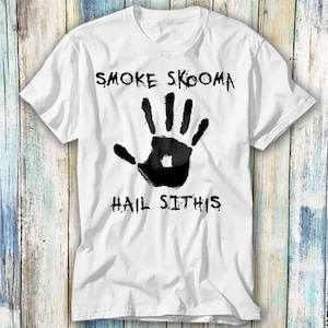 Op de afbeelding: Wit t-shirt met een zwarte handafdruk. De tekst "SMOKE SKOOMA" staat boven de handafdruk en "HAIL SITHIS" eronder. Het shirt heeft een ronde hals en korte mouwen.