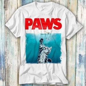 以下が含まれることがあります： 白いTシャツに、水の中から手を伸ばしている猫のグラフィックがプリントされています。猫の上に赤い文字で「PAWS」と書いてあります。