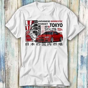 Puede incluir: Camiseta blanca con un Nissan Skyline GT-R rojo impreso en la parte delantera. El coche está rodeado de texto que dice "Japanese Domestic Market", "Tokyo", "Nissan", "GT-R", "Skyline R34" y "日本の国内市場".
