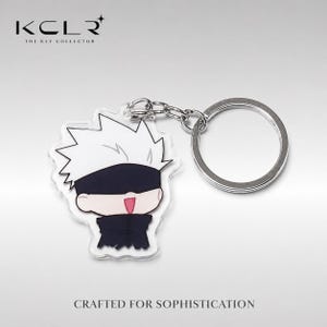 Op de afbeelding: Een acryl sleutelhanger met een chibi-stijl personage met stekelig wit haar, een zwarte blinddoek en een donkere outfit met een hoge kraag. Het personage heeft een glimlachende uitdrukking met een zichtbare roze tong. Een zilveren ring is bevestigd.
