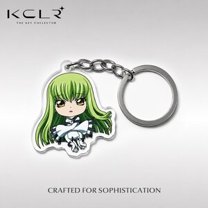 Puede incluir: Un llavero de acrílico con un personaje estilo chibi de largo cabello verde lima y ojos dorados, que viste un atuendo blanco con detalles negros. El dije está sujeto a una cadena de metal plateado y una argolla circular para llaves.