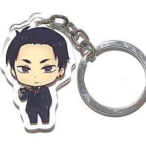 Peut inclure: Un porte-clés en acrylique transparent avec une illustration de dessin animé d'une personne aux cheveux noirs, portant un costume noir et une chemise blanche avec une cravate noire. Le porte-clés a un anneau en métal argenté.