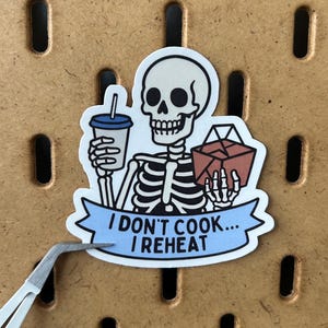 May include: A sticker featuring a cartoon skeleton holding a drink and a takeout box. The skeleton has a white skull and rib cage. The sticker has a blue banner with the text "I DON'T COOK... I REHEAT".