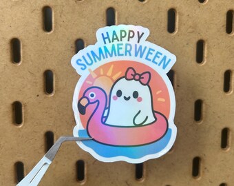 Puede incluir: Una pegatina holográfica con un fantasma de dibujos animados con un lazo flotando en un flotador de flamenco. La pegatina tiene las palabras "Happy Summerween" en un diseño azul y naranja. La pegatina está sujeta con unas pinzas.