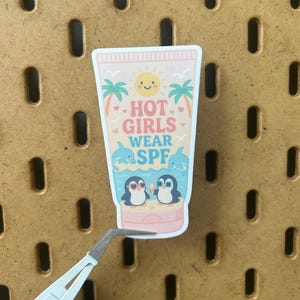 May include: A sticker in the shape of a sunscreen tube with the text "HOT GIRLS WEAR SPF". The sticker features a smiling sun, palm trees, dolphins, and two penguins wearing sunglasses.