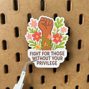 May include: A sticker featuring a brown fist surrounded by flowers and greenery. The sticker has the text "FIGHT FOR THOSE WITHOUT YOUR PRIVILEGE" in brown letters on a white background. The sticker is being held by a pair of tweezers.