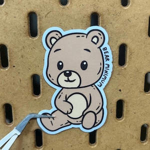 May include: A sticker featuring a cartoon teddy bear in shades of brown and beige, outlined in black, with the text "BEAR MINIMUM". The bear is sitting with its legs crossed and is being held by tweezers.