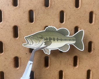 May include: A sticker of a largemouth bass fish, featuring shades of green, gray, and white. The fish is held by a pair of white tweezers against a brown pegboard background. The fish has an open mouth and detailed fins.