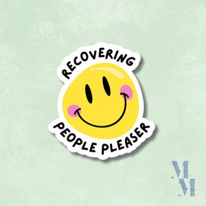 May include: A sticker with a yellow smiley face and the text "RECOVERING PEOPLE PLEASER". The smiley has black eyes and a curved black mouth, with pink blush. The sticker is on a light green background, with a white border.