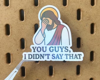 Puede incluir: Una pegatina con una ilustración de Jesús con la mano sobre los ojos. La pegatina tiene el texto "YOU GUYS, I DIDN'T SAY THAT" en azul y blanco. La pegatina está siendo sostenida por unas pinzas.
