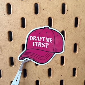 May include: A sticker of a pink baseball cap with the text "DRAFT ME FIRST" in white. The sticker is held by tweezers against a brown pegboard background.