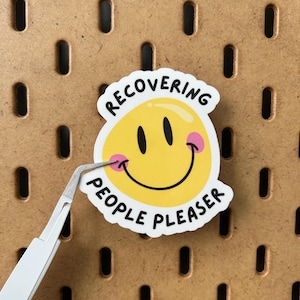 May include: A white sticker featuring a yellow smiley face with black eyes and pink cheeks. The sticker has the words "RECOVERING PEOPLE PLEASER" in black text. Tweezers are holding the sticker against a brown pegboard.
