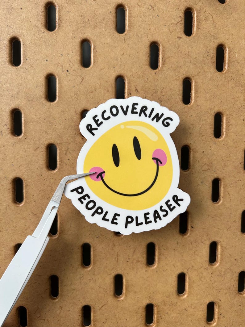 May include: A white sticker featuring a yellow smiley face with black eyes and pink cheeks. The sticker has the words "RECOVERING PEOPLE PLEASER" in black text. Tweezers are holding the sticker against a brown pegboard.