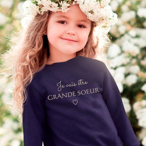 Peut inclure: Une jeune fille portant un sweat-shirt bleu marine avec le texte "Je vais être GRANDE SOEUR" et un symbole de cœur. Elle porte une couronne de fleurs blanches et une jupe en tulle rose.