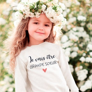 以下が含まれることがあります： 白いスウェットシャツを着た若い女の子。スウェットシャツには「Je vais être grande soeur」という文字とピンクのハートが描かれています。彼女は花冠をかぶって笑顔です。