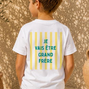 Peut inclure: Un t-shirt blanc à manches courtes avec un motif rectangulaire à rayures verticales jaunes dans le dos. Au centre des rayures, le texte "JE VAIS ÊTRE GRAND FRÈRE" est imprimé en lettres capitales bleu sarcelle. Porté avec un jean bleu.