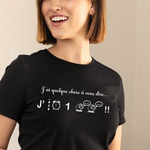Peut inclure: Un t-shirt noir avec un motif blanc qui dit "J'ai quelque chose à vous dire... J'" suivi d'une horloge, du chiffre "1" et de trois moutons avec des bulles de dialogue.