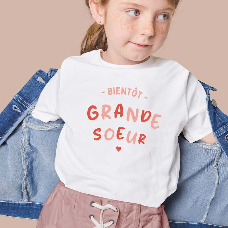 K&ouml;nnte beinhalten: Wei&szlig;es T-Shirt mit dem Text "Bient&ocirc;t - Grande Soeur" in rosa und roter Schrift mit einem kleinen rosa Herz.