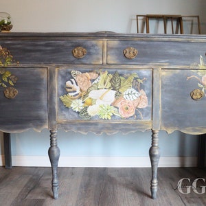 May include: A vintage, dark gray painted sideboard with gold accents and floral decals. The sideboard has three drawers and three doors. The legs are turned and have a gold finish.