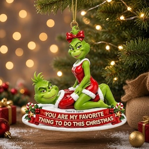 Op de afbeelding: Een kerstornament met twee groene figuren in rode outfits met witte randen. De ene figuur zit op de andere, beiden glimlachen. Een rode banner aan de onderkant bevat de tekst: Daniel & Sophia, YOU ARE MY FAVORITE THING TO DO THIS CHRISTMAS.