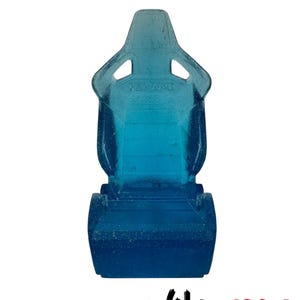 May include: A transparent blue miniature racing seat with the word "RECARO" visible. The seat has a high back with two openings near the top and a base. The logo "YAKUZIPPO" is at the bottom.